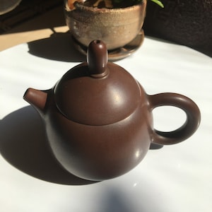 Può includere: Una teiera in ceramica marrone con un coperchio e un piccolo beccuccio. La teiera è appoggiata su una superficie bianca.