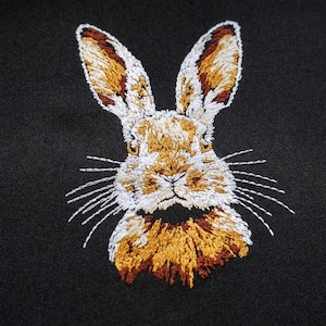 Peut inclure: Broderie repr&eacute;sentant le visage d'un lapin sur un fond de tissu noir. Le lapin a une fourrure blanche avec des accents bruns et dor&eacute;s autour des oreilles et du nez.