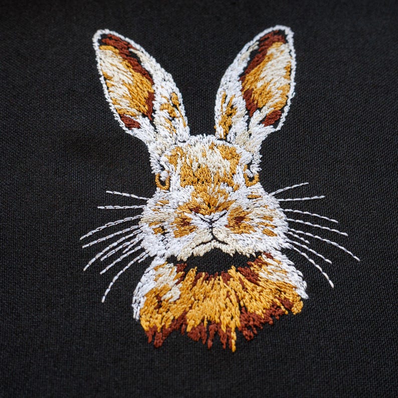 Peut inclure: Broderie repr&eacute;sentant le visage d'un lapin sur un fond de tissu noir. Le lapin a une fourrure blanche avec des accents bruns et dor&eacute;s autour des oreilles et du nez.