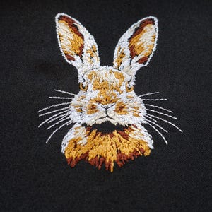 Peut inclure: Broderie de t&ecirc;te de lapin sur un fond de tissu noir. Le lapin a du poil blanc, des oreilles brunes et un nez brun.