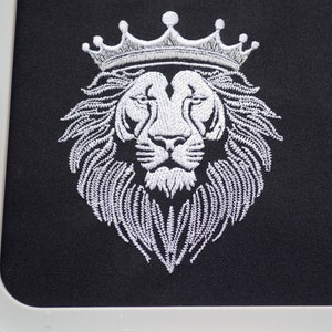 May include: White embroidered lion head wearing a crown on black fabric. The lion's mane and crown are intricately detailed. The image is framed by a white embroidery hoop, suggesting a craft project or design.