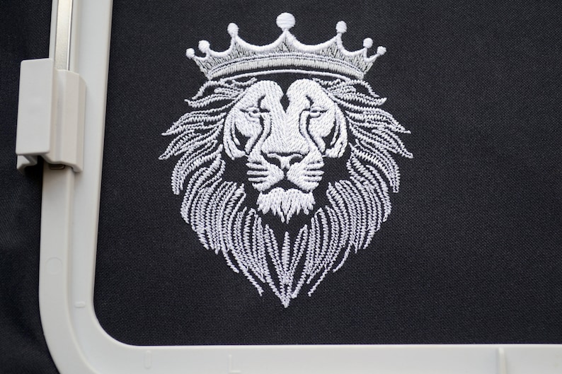 May include: White embroidered lion head wearing a crown on black fabric. The lion's mane and crown are intricately detailed. The image is framed by a white embroidery hoop, suggesting a craft project or design.