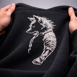 May include: Black sweatshirt featuring a detailed white embroidered wolf design. The wolf is depicted in a stylized, artistic manner, with intricate stitching creating the image. The sweatshirt is held up, showcasing the design.