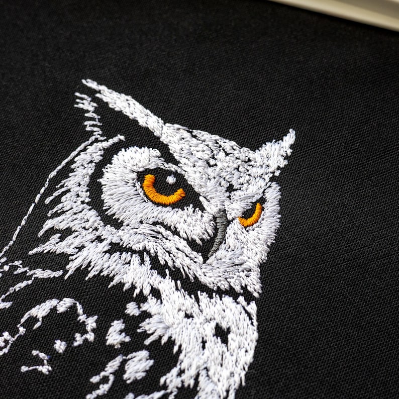 May include: Close-up of an embroidered owl on black fabric. The owl is rendered in white thread with detailed stitching, showcasing its feathers and facial features. The owl's eyes are a bright orange, creating a striking contrast against the white and black background.