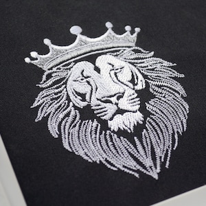 May include: A white embroidered lion's head wearing a crown on black fabric. The detailed embroidery showcases the lion's mane and facial features, creating a regal design. The image is a close-up of the embroidery.