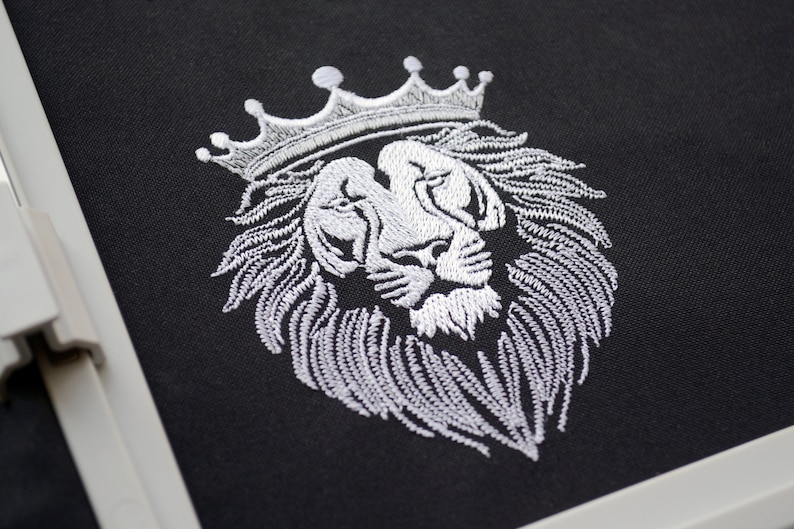 May include: A white embroidered lion's head wearing a crown on black fabric. The detailed embroidery showcases the lion's mane and facial features, creating a regal design. The image is a close-up of the embroidery.