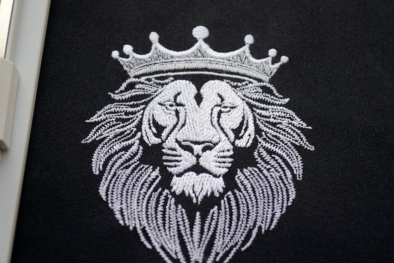 May include: White embroidered lion's head wearing a crown, set against a black backdrop. The detailed embroidery highlights the lion's mane and crown. The image is a close-up, emphasizing the texture and design of the embroidery.