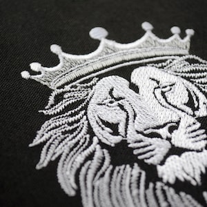 May include: Close-up of black fabric featuring a white embroidered lion's head wearing a crown. The embroidery shows detailed stitching, emphasizing the lion's mane and facial features. The crown is positioned on the lion's head.