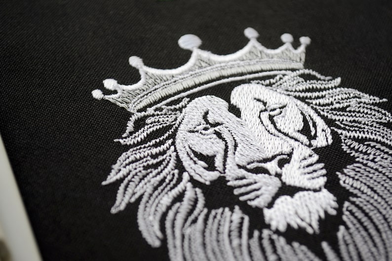 May include: Close-up of black fabric featuring a white embroidered lion's head wearing a crown. The embroidery shows detailed stitching, emphasizing the lion's mane and facial features. The crown is positioned on the lion's head.