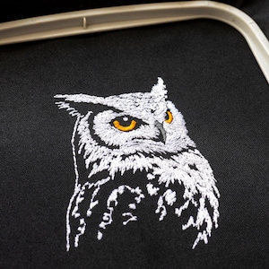 May include: Embroidered owl design on black fabric. The owl is depicted in white thread with yellow eyes, showcasing detailed feathering. The image is framed by an embroidery hoop, suggesting a craft project.
