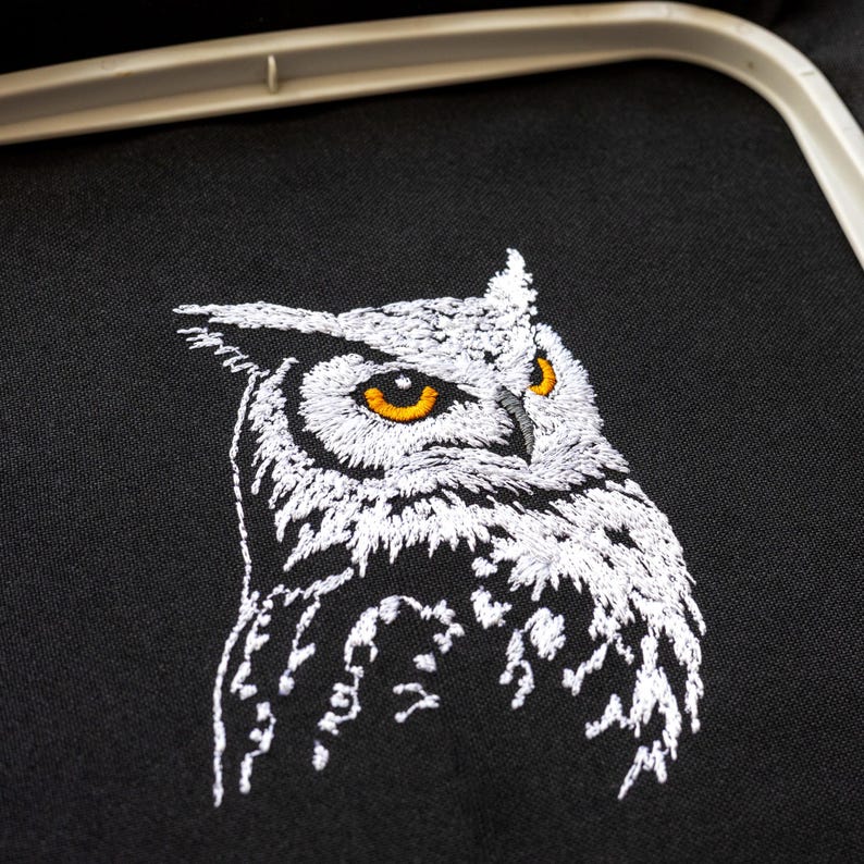 May include: Embroidered owl design on black fabric. The owl is depicted in white thread with yellow eyes, showcasing detailed feathering. The image is framed by an embroidery hoop, suggesting a craft project.