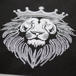 May include: White embroidered lion head with a crown on black fabric. The lion's mane and crown are intricately detailed, creating a regal and striking design. The embroidery is clean and precise, highlighting the lion's features.