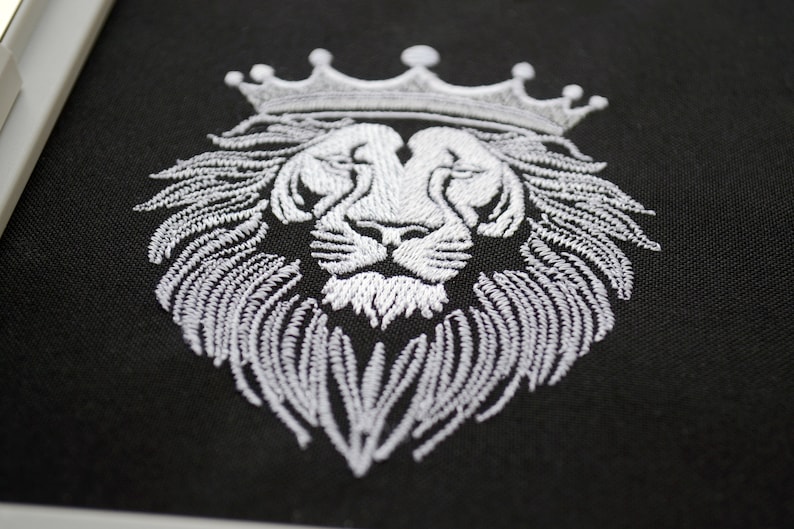 May include: White embroidered lion head with a crown on black fabric. The lion's mane and crown are intricately detailed, creating a regal and striking design. The embroidery is clean and precise, highlighting the lion's features.