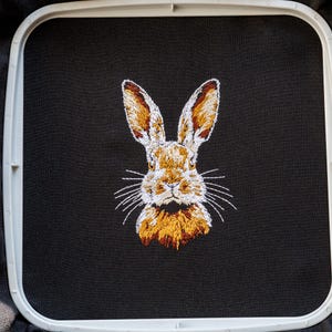 Peut inclure: Image brod&eacute;e d'un visage de lapin sur un tissu noir. Le lapin a une fourrure brune, des moustaches blanches et de la fourrure blanche autour du nez.