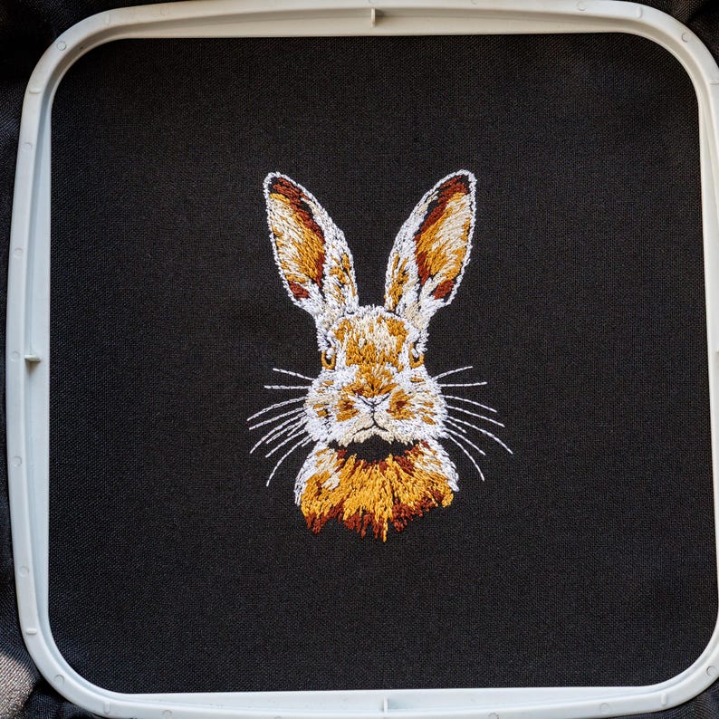Peut inclure: Image brod&eacute;e d'un visage de lapin sur un tissu noir. Le lapin a une fourrure brune, des moustaches blanches et de la fourrure blanche autour du nez.