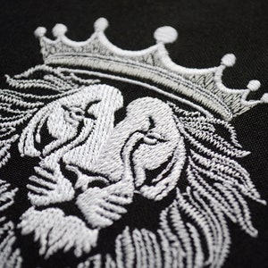May include: A detailed white embroidered lion's head with a crown, set against a black backdrop. The embroidery showcases the lion's mane, face, and crown. This design is suitable for clothing or home decoration.