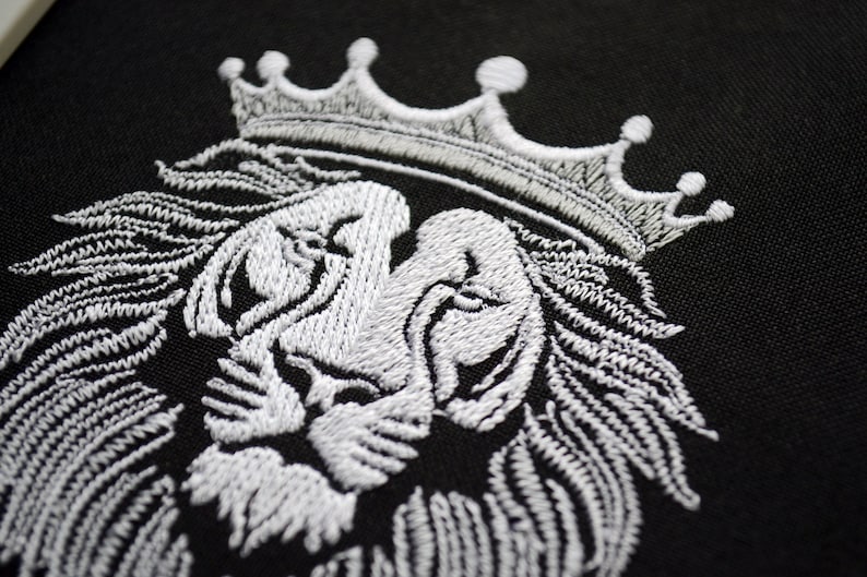 May include: A detailed white embroidered lion's head with a crown, set against a black backdrop. The embroidery showcases the lion's mane, face, and crown. This design is suitable for clothing or home decoration.