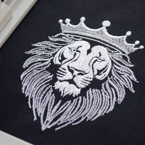 May include: White embroidered lion head wearing a crown on black fabric. The detailed embroidery shows the lion's mane and facial features. The design is suitable for clothing or home decor.