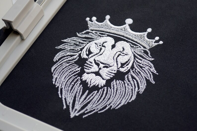 May include: White embroidered lion head wearing a crown on black fabric. The detailed embroidery shows the lion's mane and facial features. The design is suitable for clothing or home decor.