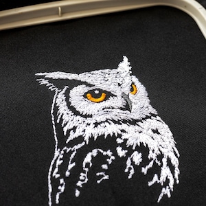 May include: Close-up of an embroidered owl on black fabric. The owl is white with detailed stitching, featuring bright orange eyes. The embroidery is set within a white hoop, showcasing a detailed craft project.