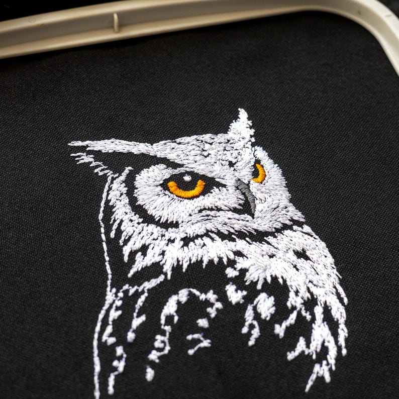 May include: Close-up of an embroidered owl on black fabric. The owl is white with detailed stitching, featuring bright orange eyes. The embroidery is set within a white hoop, showcasing a detailed craft project.