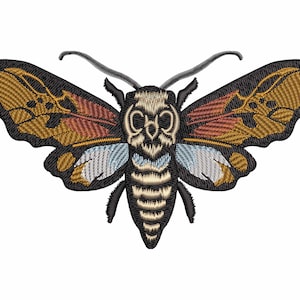 Könnte beinhalten: Gestickter Aufnäher eines schwarzen und braunen Falters mit einem schädelartigen Gesicht. Der Falter hat ein hellblaues und goldenes Muster auf seinen Flügeln.