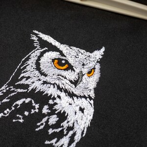May include: Embroidered owl design on black fabric. The owl features white thread detailing with orange eyes. The artwork is a close-up of the owl's head and upper body, showcasing the intricate stitching and texture of the embroidery.