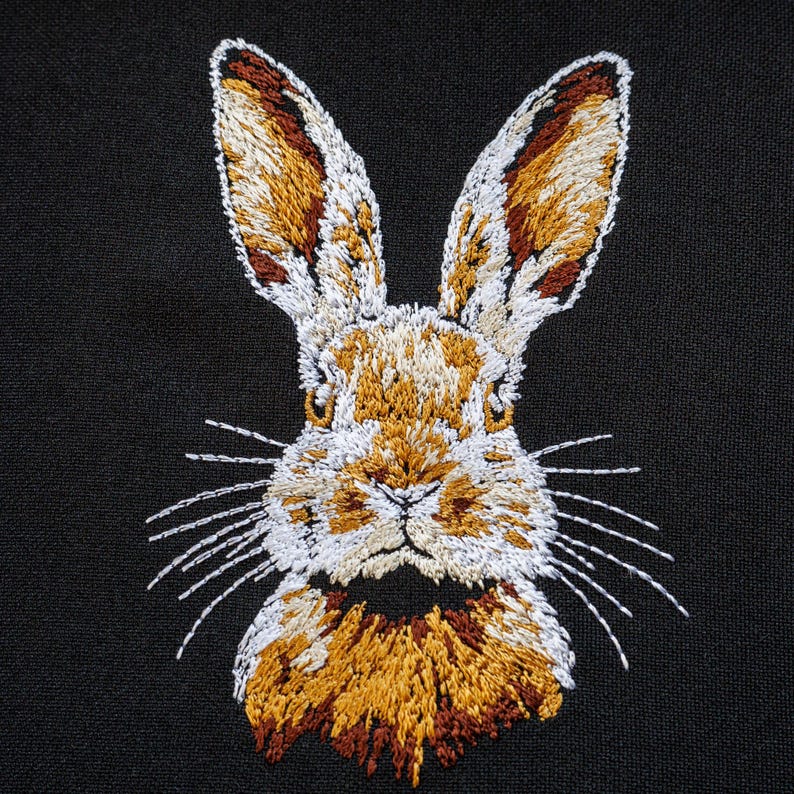 Peut inclure: Broderie repr&eacute;sentant le visage d'un lapin sur un fond de tissu noir. Le lapin a une fourrure blanche avec des accents bruns et beiges sur ses oreilles et autour de son nez.
