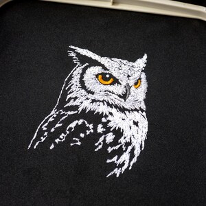 May include: Embroidered owl design on black fabric. The owl is depicted in white thread with yellow eyes, showcasing detailed stitching. The artwork is set within an embroidery hoop, suggesting a handmade craft project or finished product.
