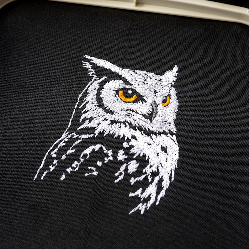 May include: Embroidered owl design on black fabric. The owl is depicted in white thread with yellow eyes, showcasing detailed stitching. The artwork is set within an embroidery hoop, suggesting a handmade craft project or finished product.