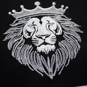 May include: Embroidered white lion's head wearing a crown, set against a black backdrop. The lion's mane and crown are intricately stitched. A close-up view of the embroidery.
