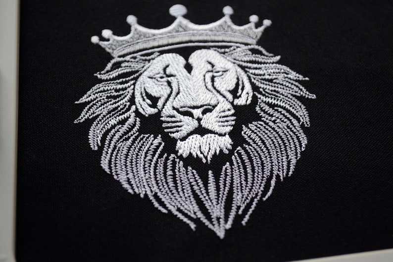 May include: Embroidered white lion's head wearing a crown, set against a black backdrop. The lion's mane and crown are intricately stitched. A close-up view of the embroidery.