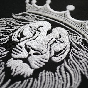 May include: Close-up of a white embroidered lion's head wearing a crown, set against a black backdrop. The intricate stitching highlights the lion's mane and facial details. This design is suitable for clothing and accessories.