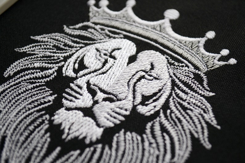 May include: Close-up of a white embroidered lion's head wearing a crown, set against a black backdrop. The intricate stitching highlights the lion's mane and facial details. This design is suitable for clothing and accessories.