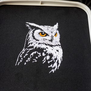 May include: Embroidered owl design on black fabric. The owl is depicted in white thread with striking orange eyes. The embroidery is set within a white hoop, showcasing the detailed craftsmanship of the artwork.