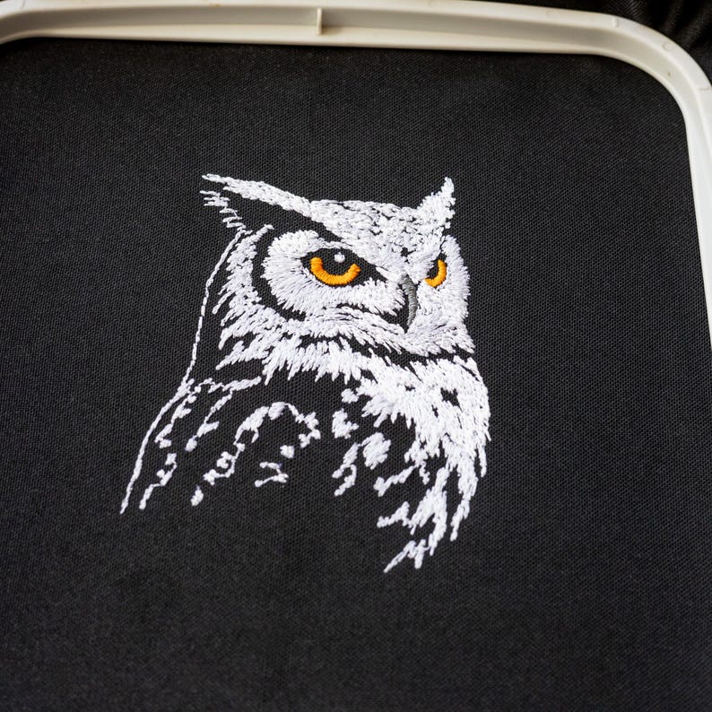 May include: Embroidered owl design on black fabric. The owl is depicted in white thread with striking orange eyes. The embroidery is set within a white hoop, showcasing the detailed craftsmanship of the artwork.