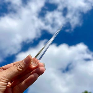 Puede incluir: Una varita de cristal transparente y puntiaguda se sostiene contra un cielo azul brillante con nubes blancas. El cristal es transparente y se estrecha hasta un punto afilado, reflejando la luz. La varita se sostiene en una mano.