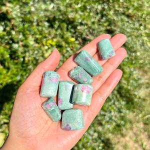 May include: A selection of tumbled ruby in zoisite stones, each showing a blend of green and pink colours. The stones are held in an open hand, with a blurred green backdrop.