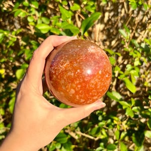 Könnte beinhalten: Eine große, polierte, rote Achatkugel mit weißen und braunen Einschlüssen. Die Kugel wird in einer Hand gehalten.