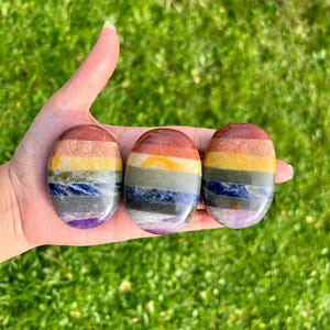 Könnte beinhalten: Drei ovale Chakra-Steine mit einem geschichteten Design in verschiedenen Farben, darunter Rot, Orange, Gelb, Grün, Blau und Lila. Die Steine werden in einer Hand vor einem Hintergrund aus grünem Gras gehalten.