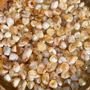 May include: A bowl filled with tumbled citrine stones. The stones are a light yellow colour with some having a darker, more golden hue. The stones are smooth and polished.