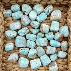 Op de afbeelding: Een verzameling getrommelde blauwe en bruine Larimar-stenen in een geweven mand. De stenen zijn glad en hebben verschillende patronen.