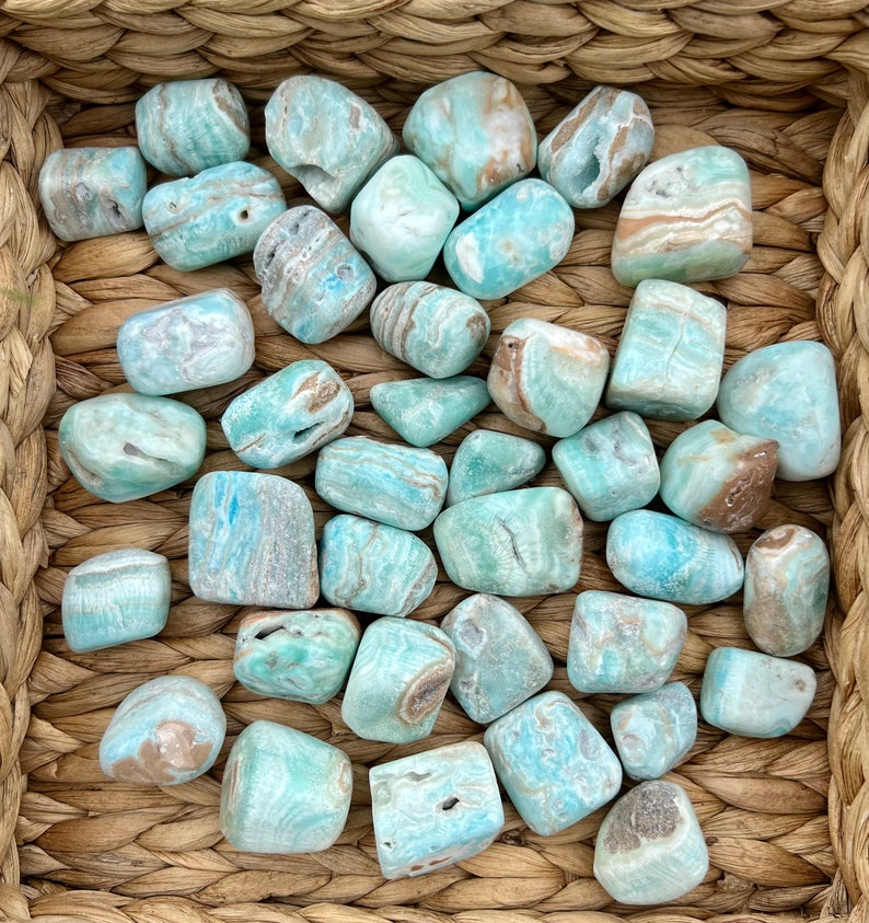 Op de afbeelding: Een verzameling getrommelde blauwe en bruine Larimar-stenen in een geweven mand. De stenen zijn glad en hebben verschillende patronen.