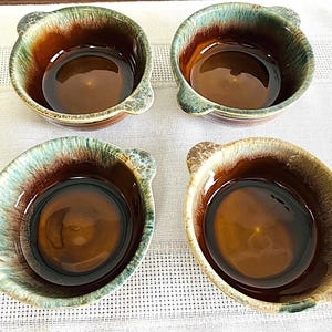 May include: Four ceramic bowls with a brown interior and a two-tone exterior. The exterior features a blend of brown, green, and blue hues. Each bowl has two small handles. The bowls are arranged on a white surface.