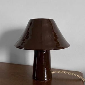 Może przedstawiać: Brązowa ceramiczna lampa stołowa w kształcie grzyba. Lampa ma stożkowy klosz i cylindryczny trzon. Lampa stoi na drewnianej powierzchni. Brązowy przewód wychodzi z podstawy.