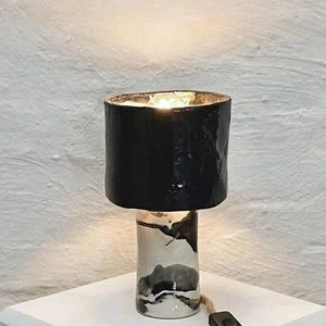 Puede incluir: Lámpara de mesa de cerámica con pantalla negra y base cilíndrica. La base presenta un diseño veteado en blanco y negro. La lámpara tiene un cable con un interruptor de encendido/apagado. La lámpara está sobre una superficie blanca.