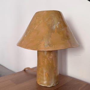 May include: A ceramic mushroom-shaped table lamp with a warm, earthy color palette. The lamp features a conical shade and a cylindrical base, with a textured surface. The lamp is sitting on a wooden surface.