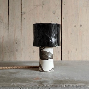 May include: A ceramic table lamp with a black lampshade and a white and gray patterned base. The lamp sits on a concrete surface with a woven cord. The background is a light-colored wooden wall.