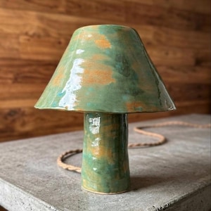 May include: A ceramic table lamp with a green and orange glazed finish. The lamp has a conical shade and a cylindrical base. The lamp is sitting on a gray surface with a wooden background. A braided cord extends from the base.