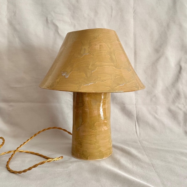 May include: A mushroom-shaped table lamp with a glossy, mustard-yellow ceramic finish featuring subtle, swirling tonal variations. The lamp includes a cylindrical base and a wide, conical shade, accompanied by a twisted gold-coloured power cord.